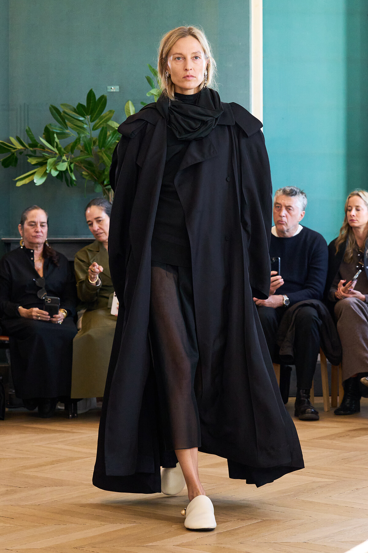Model walks a fashion show wearing a long black layered cape over black outfit with white slides; audience seated in background.