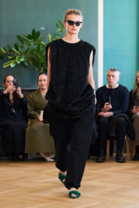 Model walks a runway in a textured black sleeveless top, loose black pants, and green sandals, wearing sunglasses as spectators watch.