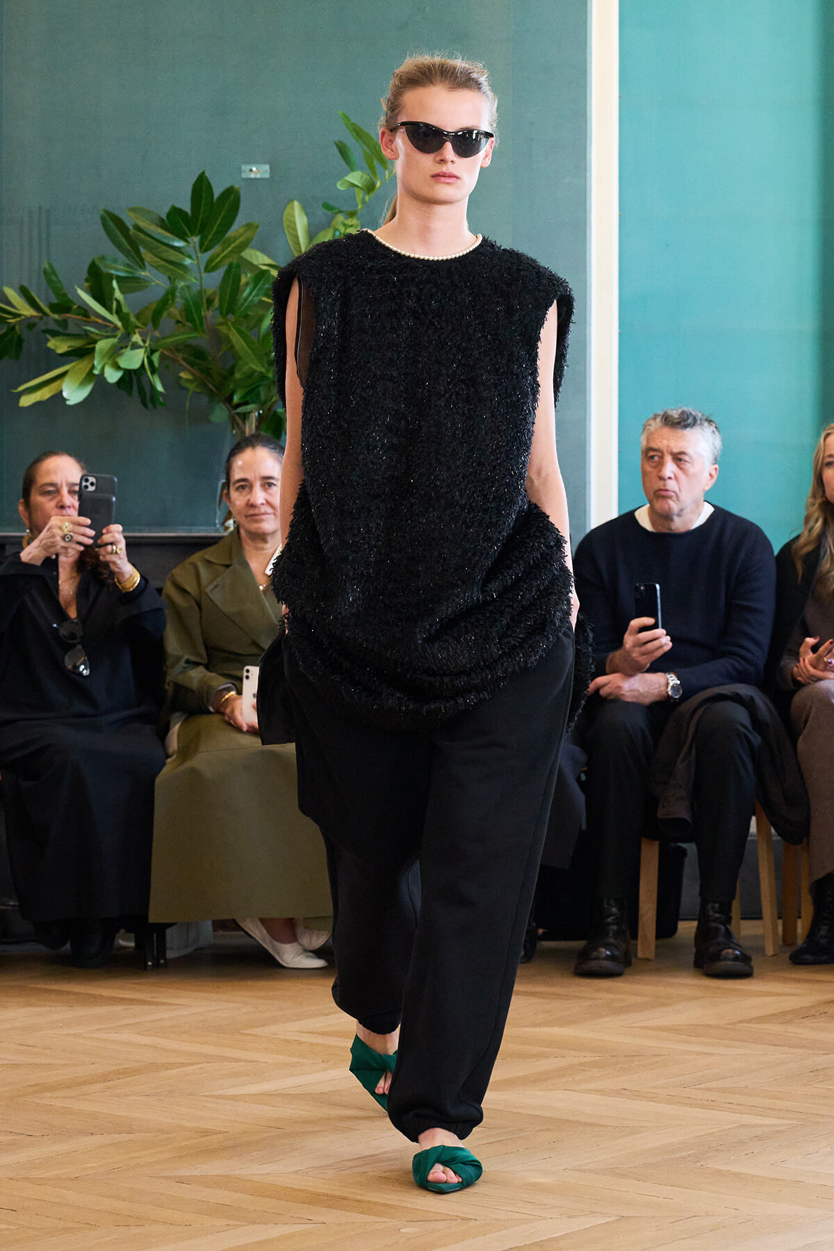 Model walks a runway in a textured black sleeveless top, loose black pants, and green sandals, wearing sunglasses as spectators watch.