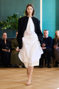 Model walks a runway in a loose white dress with a black cape-like jacket; seated audience watches in the background.