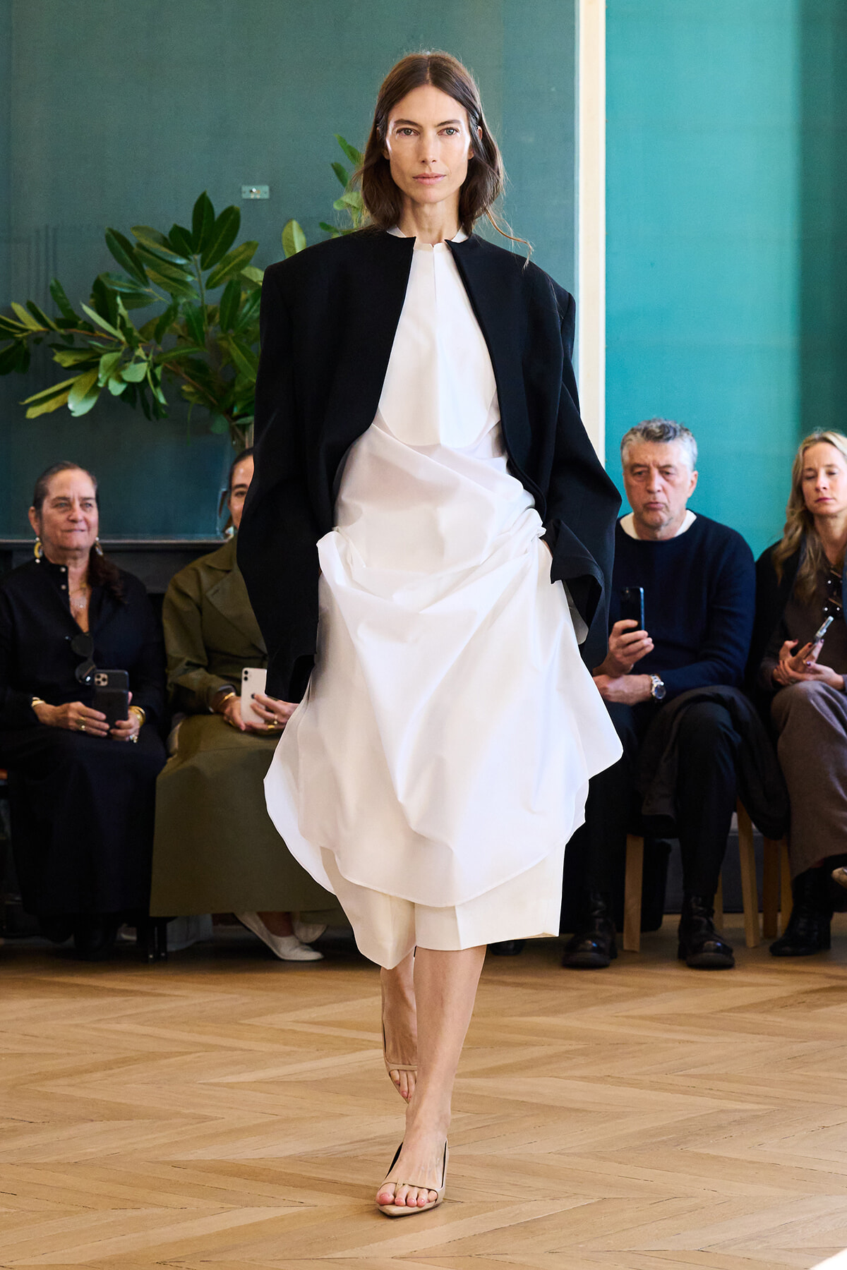 Model walks a runway in a loose white dress with a black cape-like jacket; seated audience watches in the background.