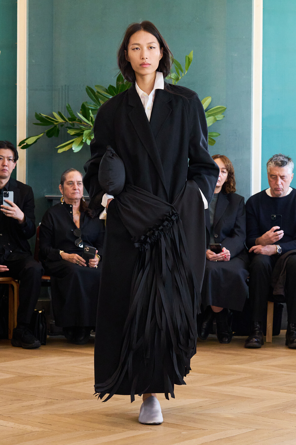 Model walks a fashion show in a long black overcoat with fringe details, white shirt collar visible, on a wooden floor with seated audience behind.