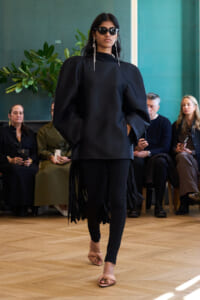 Model walks a runway in an all-black outfit with voluminous sleeves and fringe panels, wearing sunglasses and long earrings, audience in the background.