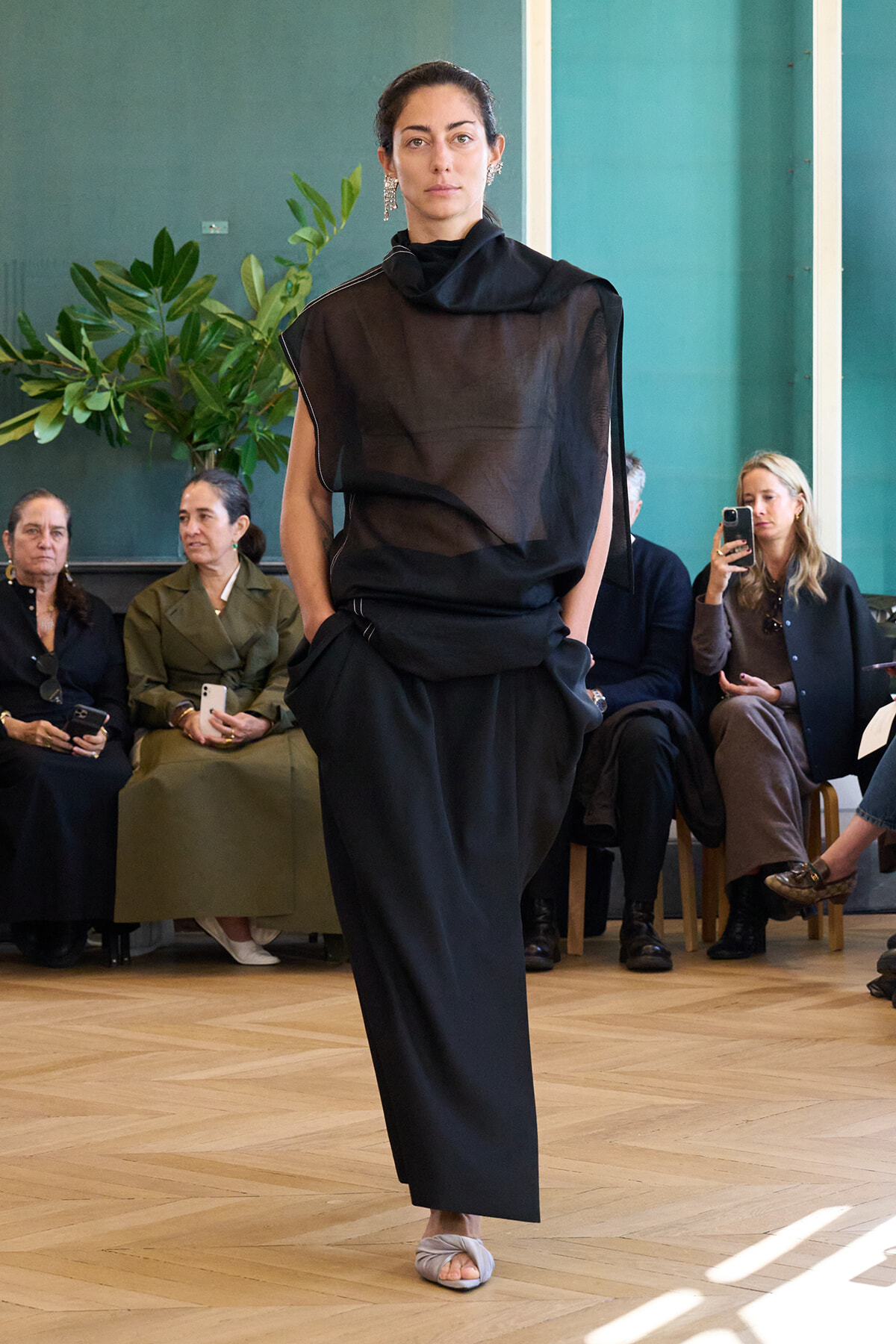 Model in a black sheer top and loose black pants walks down a wooden runway; audience sits along the sides, with plants and a teal wall in the background.