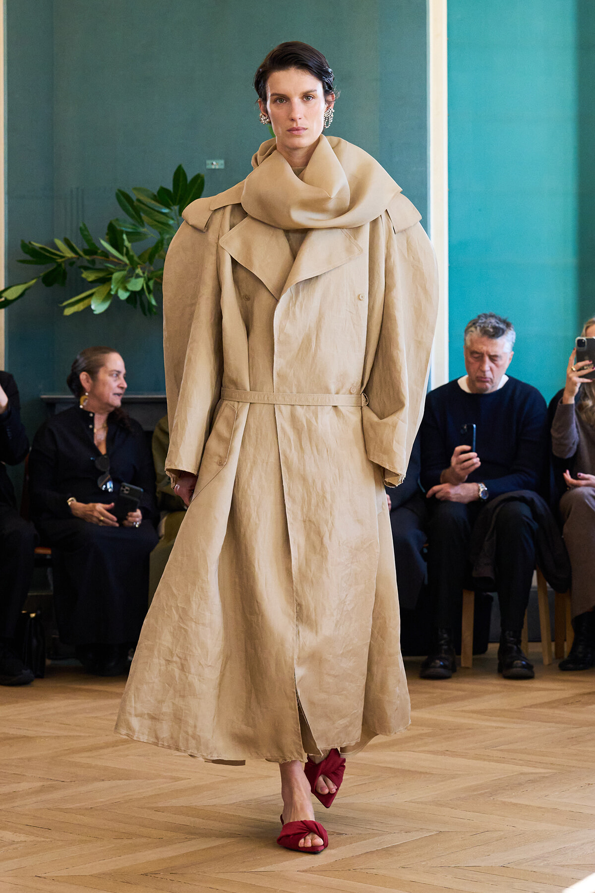 Model walks the runway in a long beige trench with a large draped collar and belt; red sandals, audience seated behind.