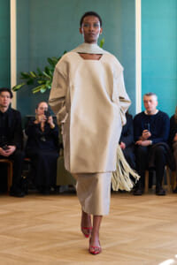 Model walks a runway in a loose beige ensemble with fringe, pink heels, and an audience in the background.