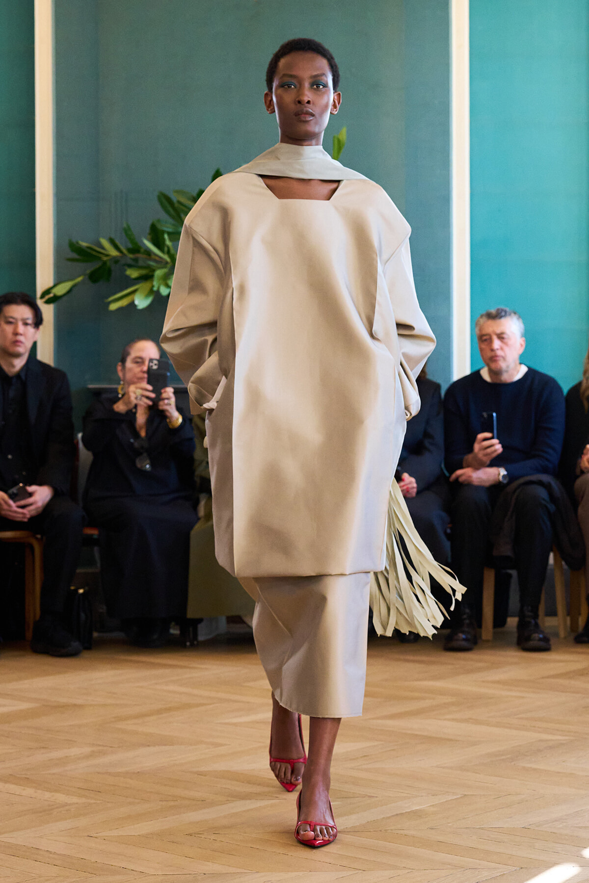 Model walks a runway in a loose beige ensemble with fringe, pink heels, and an audience in the background.