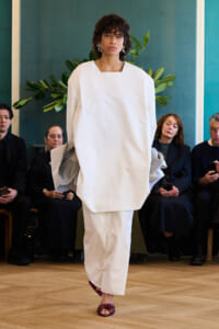 Model walks a runway in a loose white, square-neck outfit with wide sleeves; audience seated along teal wall backdrop, wearing dark attire and watching.