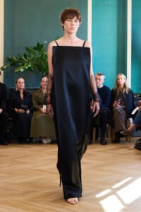 Model walking the runway in a black off-shoulder dress with sheer long sleeves, audience seated in a bright room behind her
