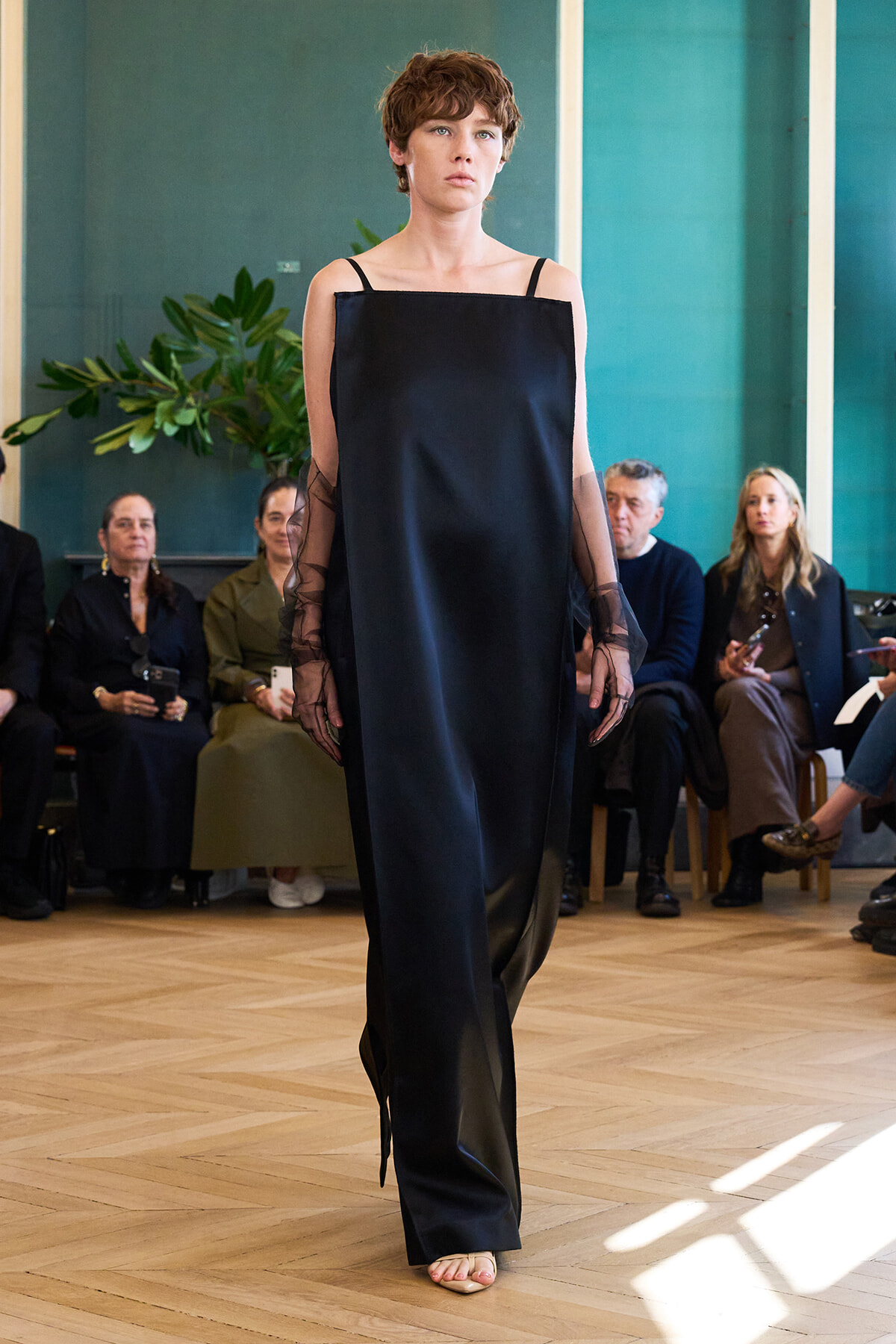 Model walking the runway in a black off-shoulder dress with sheer long sleeves, audience seated in a bright room behind her