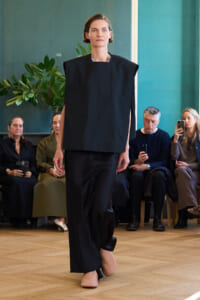 Model walks a runway in a black oversized top and wide-leg pants while seated audience watches, indoors with teal walls and plants in the background.