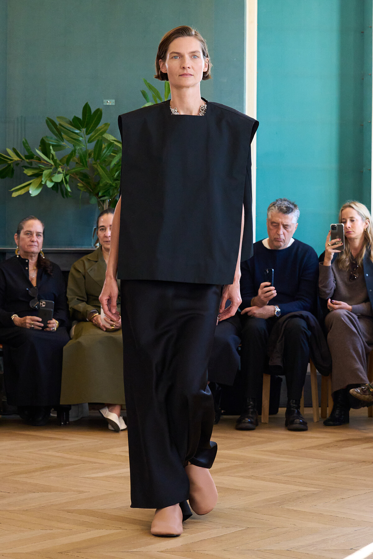Model walks a runway in a black oversized top and wide-leg pants while seated audience watches, indoors with teal walls and plants in the background.