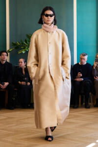 Model walks the runway in a long beige coat, pink scarf, black sunglasses, and statement earrings; audience seated in the background.