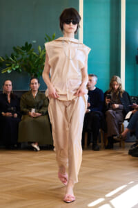 Model walking a runway in a pale beige sleeveless top and matching pants, pink twisted sandals, and sunglasses with seated spectators in the background.