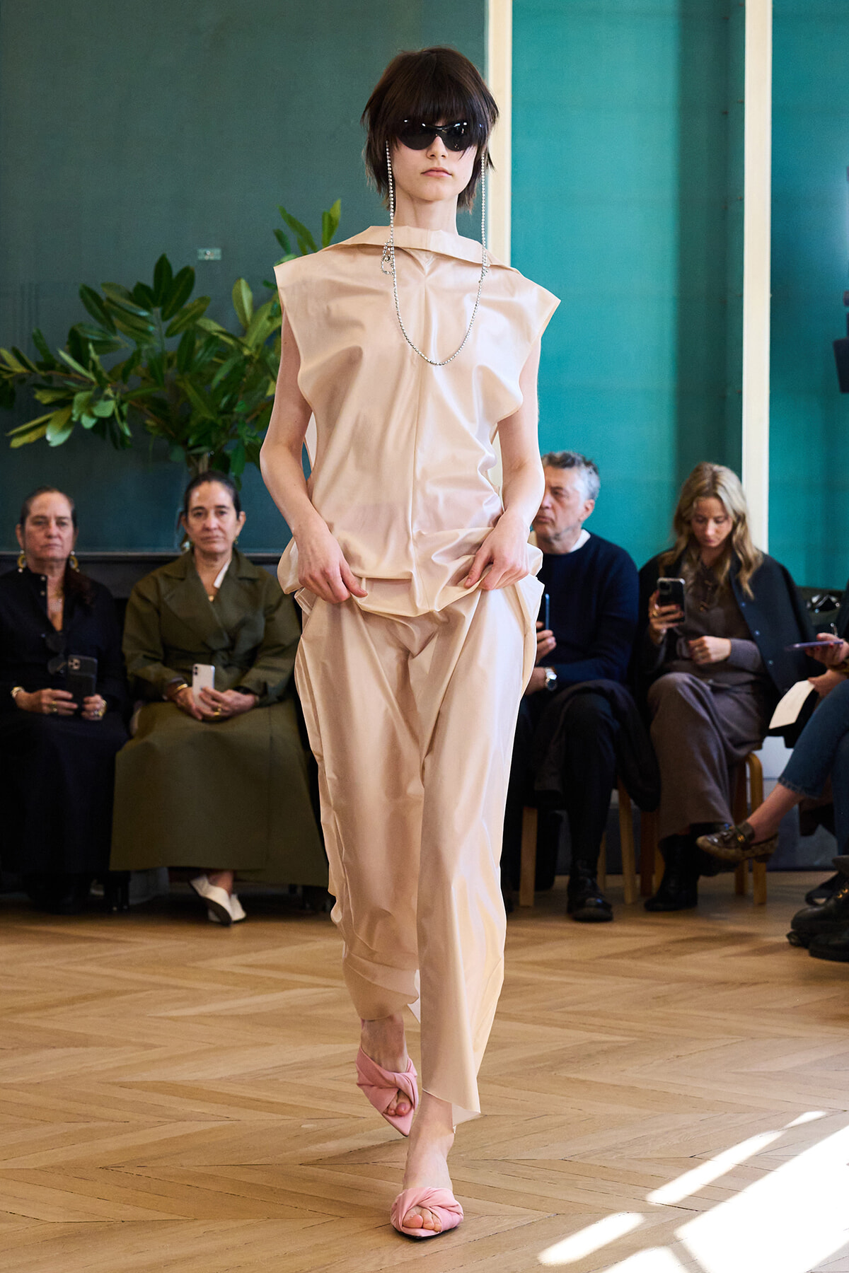 Model walking a runway in a pale beige sleeveless top and matching pants, pink twisted sandals, and sunglasses with seated spectators in the background.