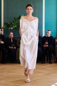 Model walks a runway in a loose, off-shoulder white dress with geometric draping; audience seated in background.