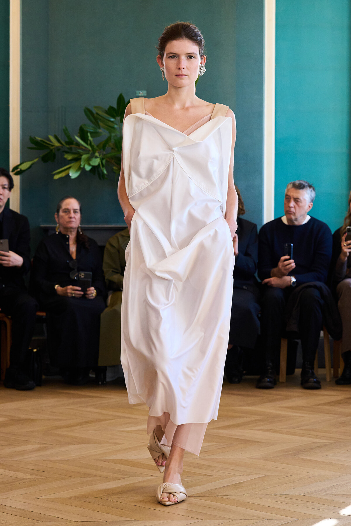 Model walks a runway in a loose, off-shoulder white dress with geometric draping; audience seated in background.