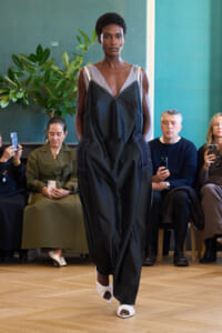 Model walks a runway in a loose black jumpsuit with a gray V-neck top and white wrapped sandals; audience watches in the background.