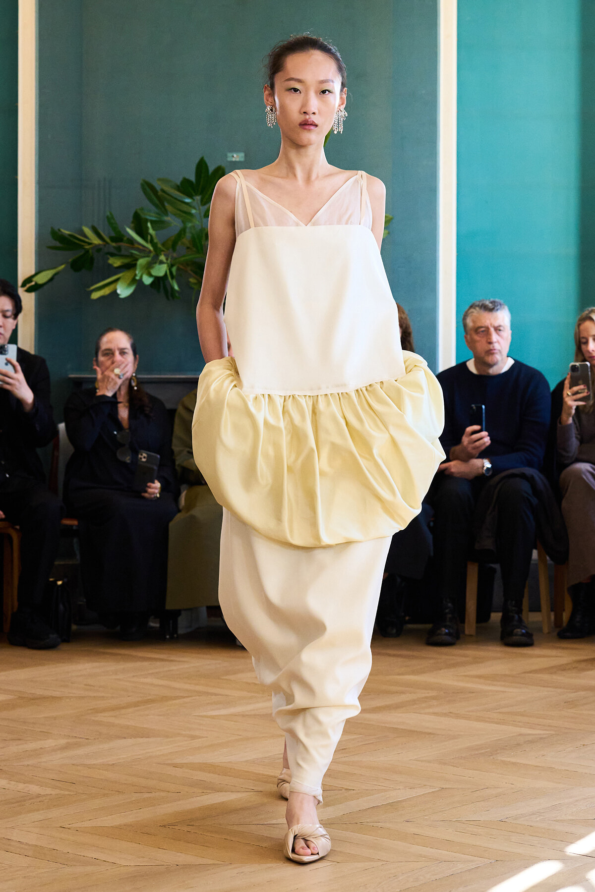 Model on a runway wearing a cream layered gown with a voluminous balloon skirt and sheer top, paired with chandelier earrings; audience watches in the background.