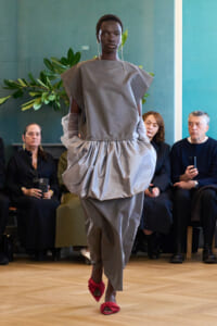 Model walks a runway in a voluminous gray dress with a structured top and gathered waist, paired with red twisted sandals as bystanders watch.