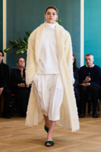 Model walks runway in white high-neck outfit with a long cream fluffy coat, green sandals, audience watching in background