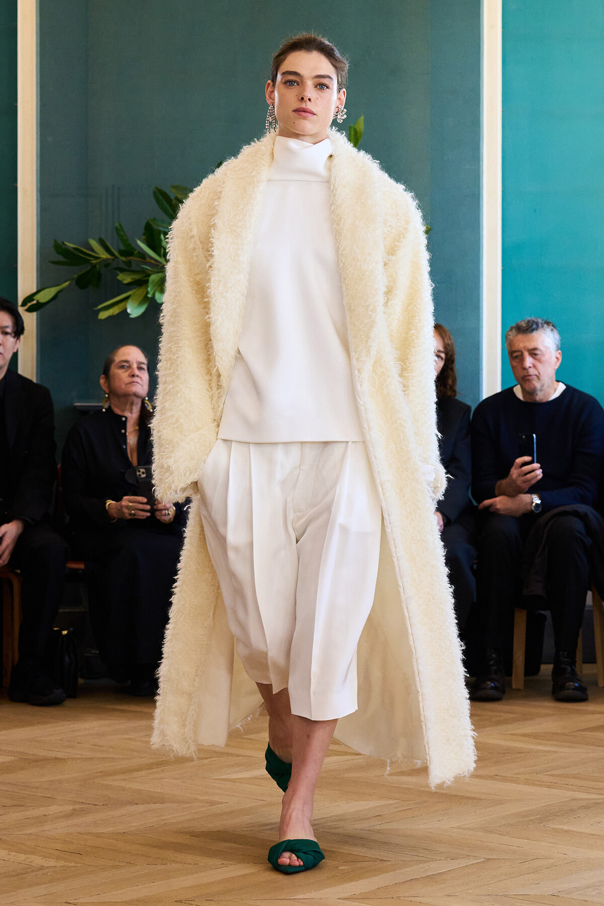 Model walks runway in white high-neck outfit with a long cream fluffy coat, green sandals, audience watching in background