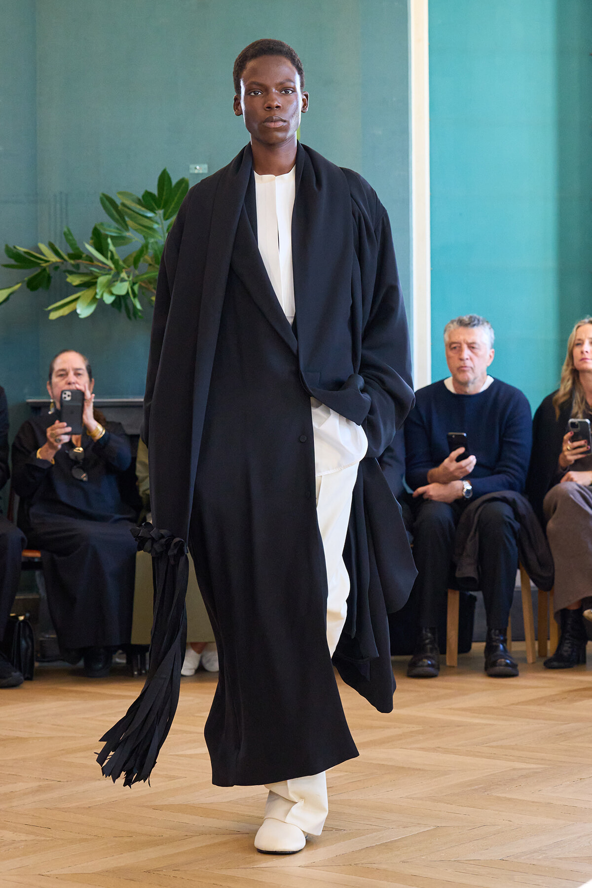 Model walking a runway in an oversized black coat over a white outfit, fringe tassels at the sleeve, with seated spectators in a teal-walled room.