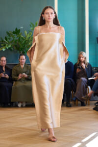 Model walking a runway in a cream satin off-shoulder dress with cape-like sleeves, audience watching the show.