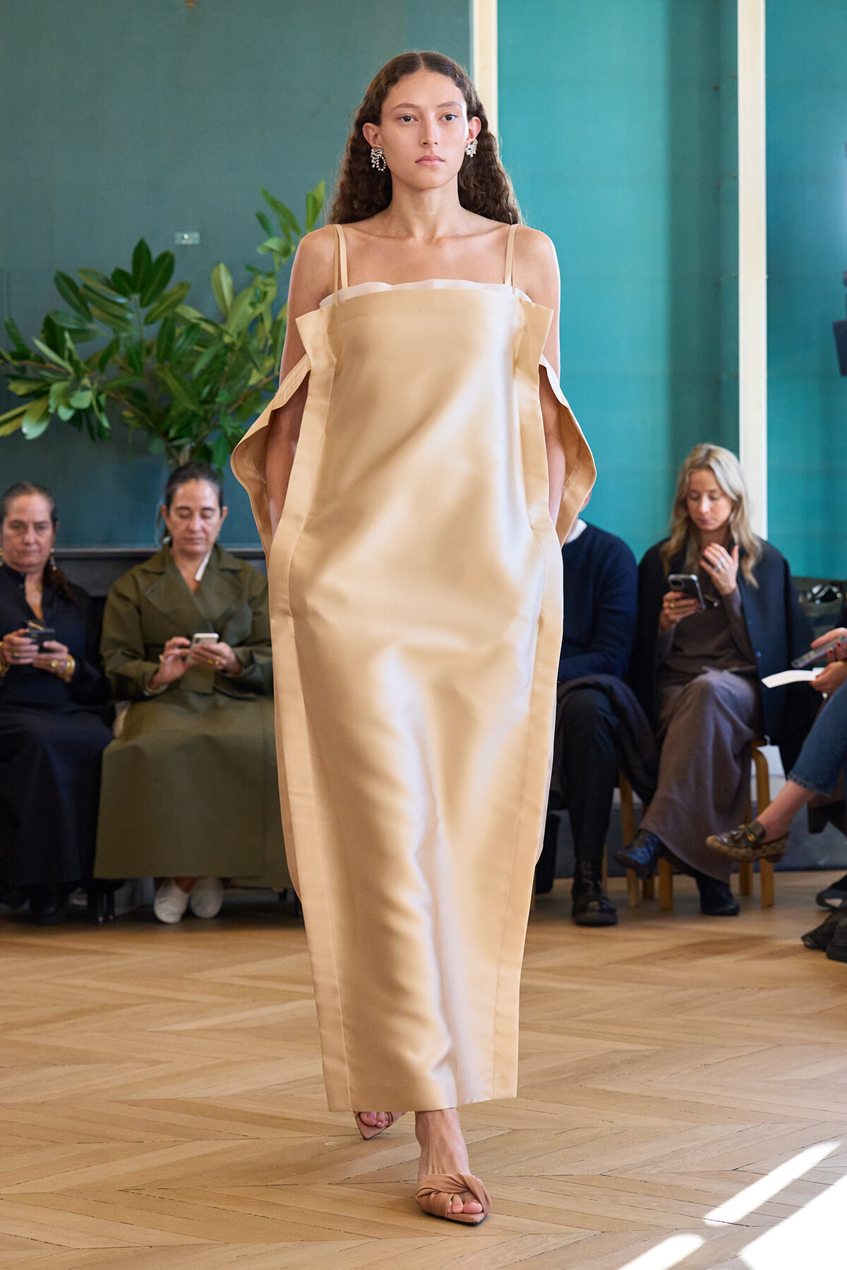 Model walking a runway in a cream satin off-shoulder dress with cape-like sleeves, audience watching the show.