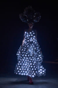 Model walks in a dark setting wearing a floor-length gown made of glowing circular embellishments and a large feathered headpiece, creating a dramatic, illuminated silhouette.