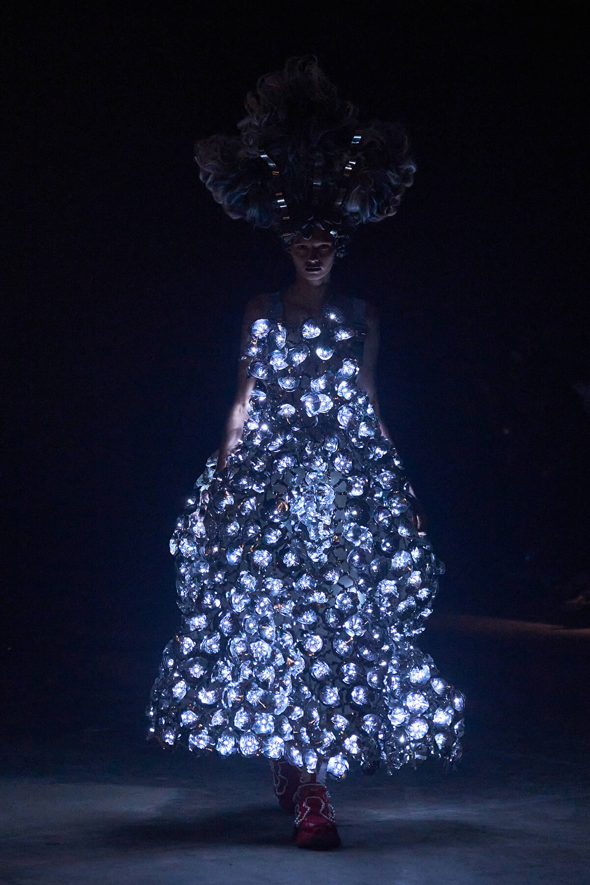 Model walks in a dark setting wearing a floor-length gown made of glowing circular embellishments and a large feathered headpiece, creating a dramatic, illuminated silhouette.