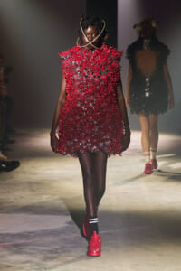 Runway model wearing a bright red, spiky-textured mini dress with a sculptural gold neckpiece and red sneakers, walking the catwalk.