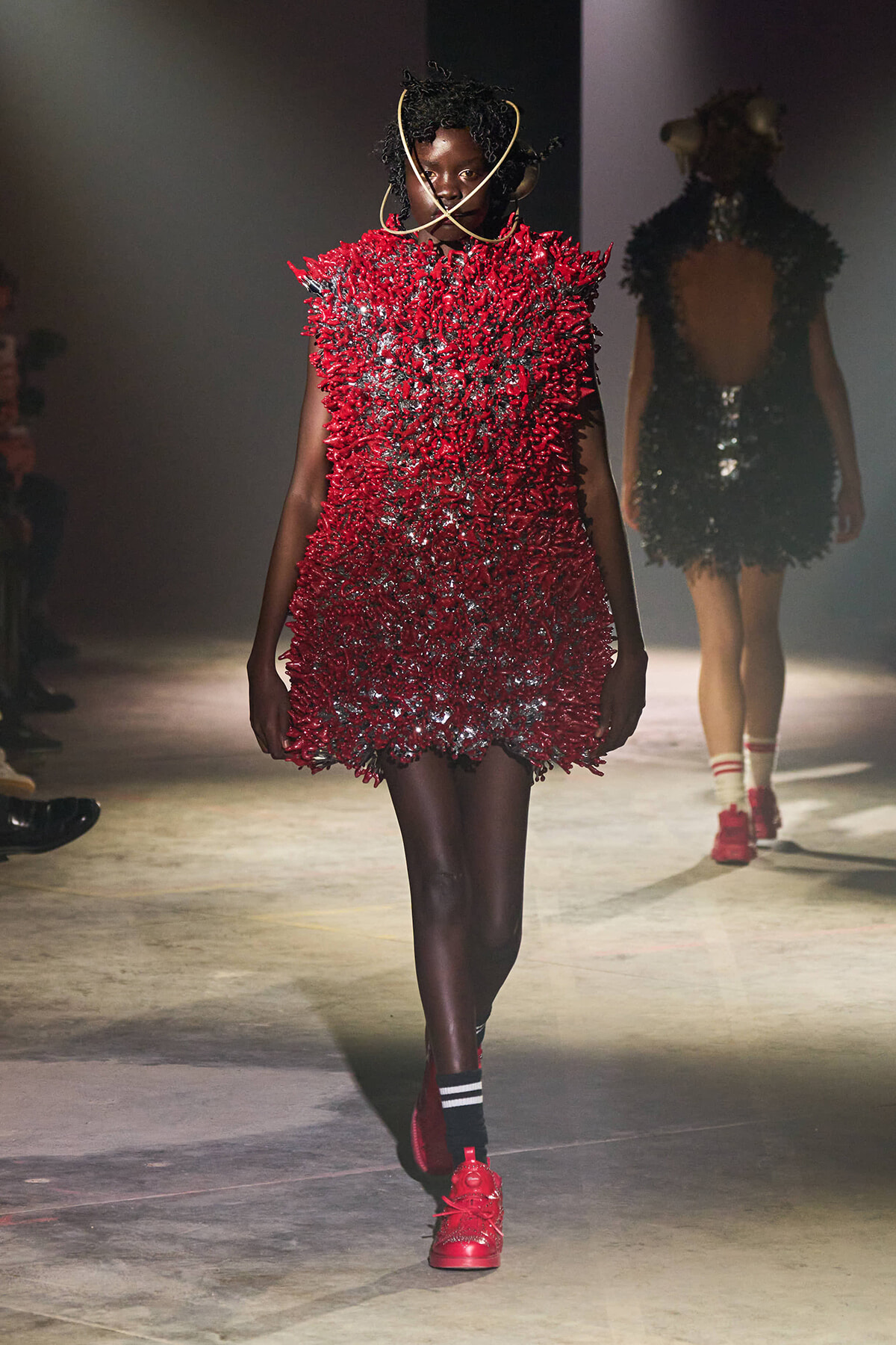 Runway model wearing a bright red, spiky-textured mini dress with a sculptural gold neckpiece and red sneakers, walking the catwalk.