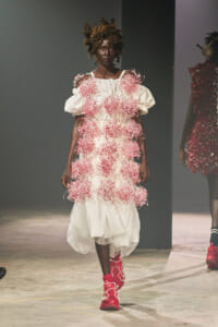 Model walks a runway in a white dress adorned with pink beaded clusters and voluminous sleeves, paired with bright pink boots.