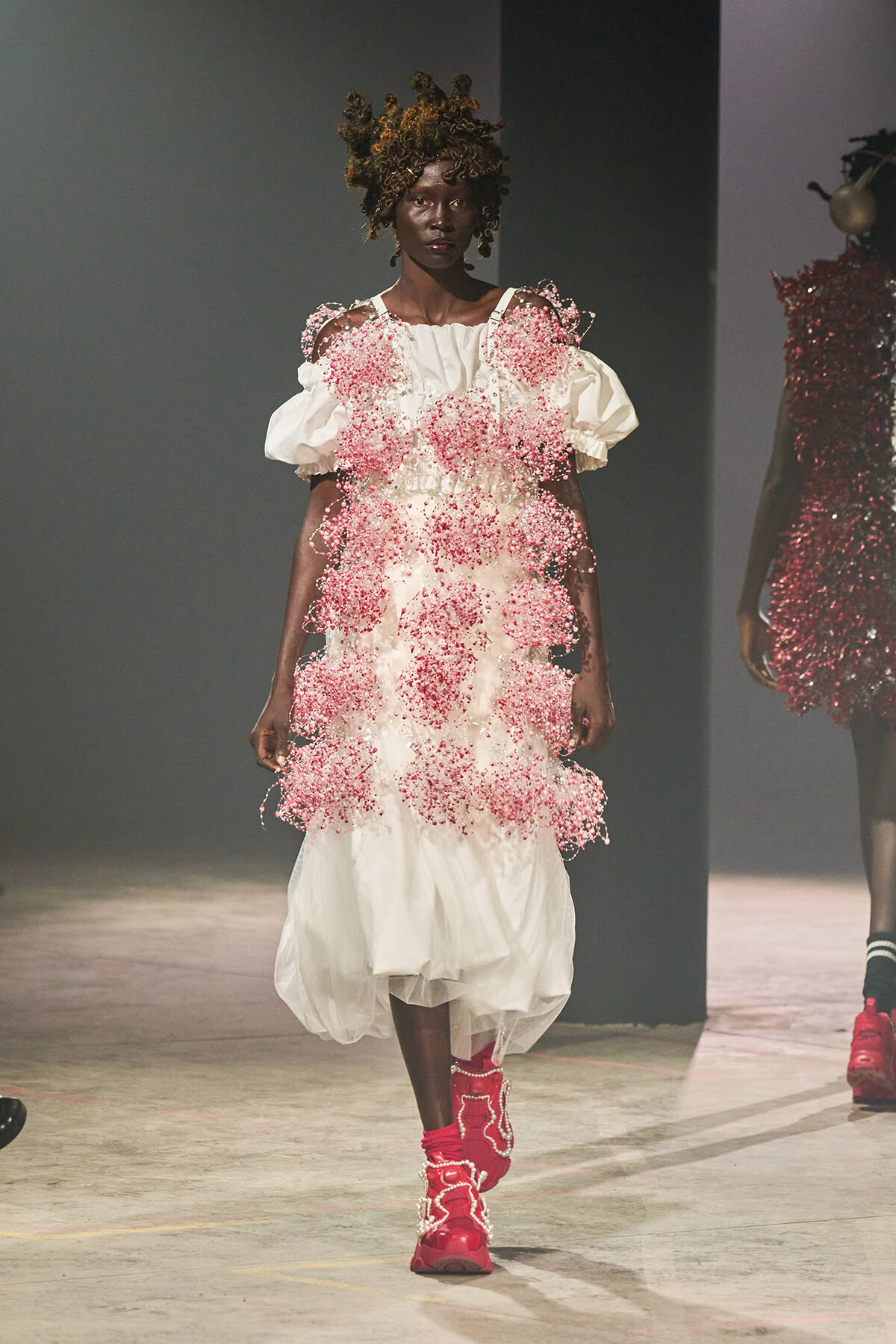 Model walks a runway in a white dress adorned with pink beaded clusters and voluminous sleeves, paired with bright pink boots.