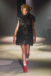 Model walks the runway in a black, spiky-textured mini dress with a gold sculptural headpiece and bright red sneakers with white socks.
