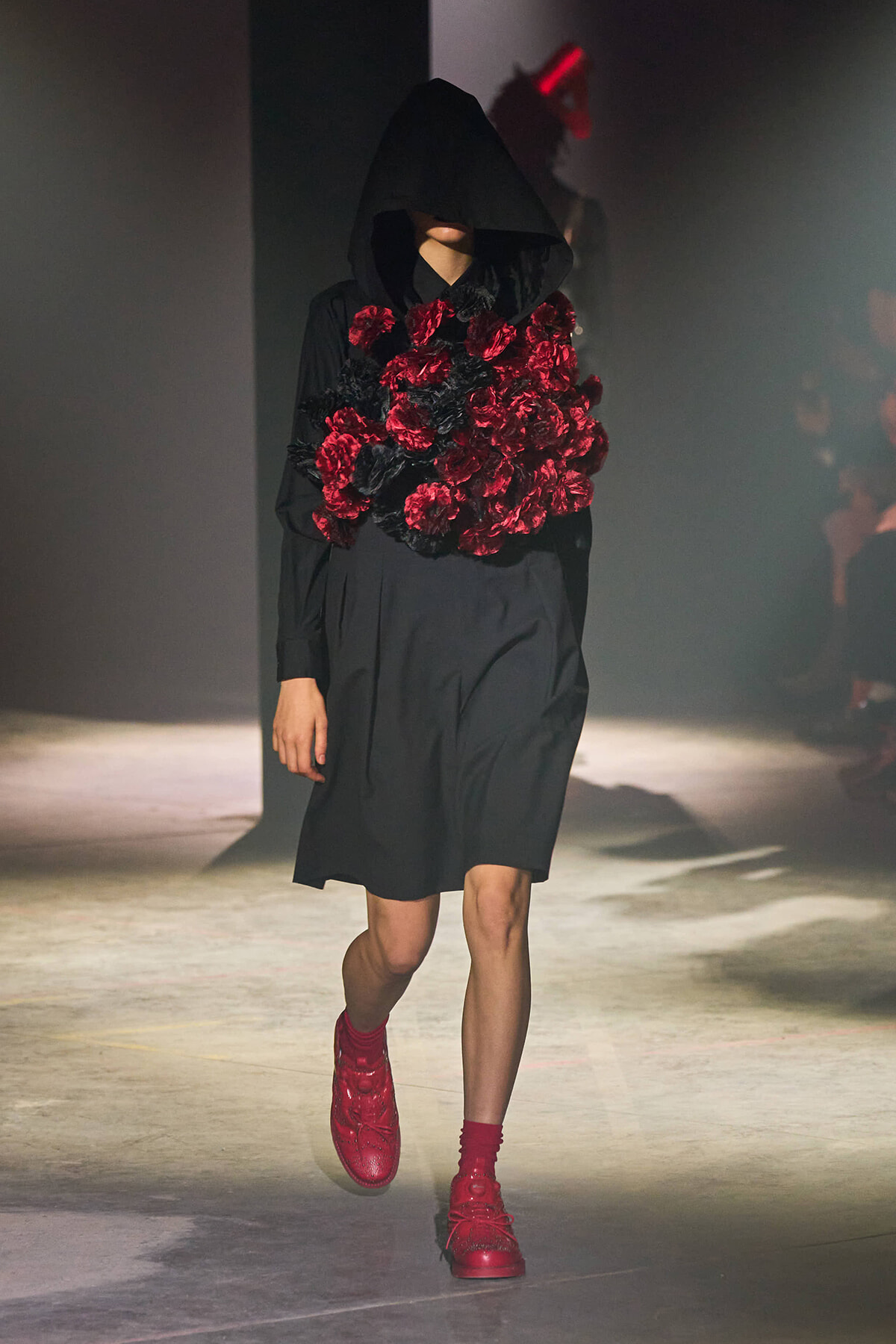 Model in a black hooded coat walking down a runway, with a dense cluster of red and black flowers around the chest and red boots visible.