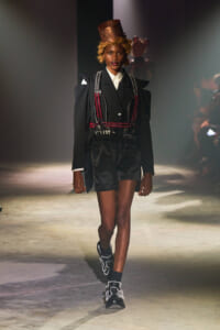 Model walks a fashion runway in a black tailored suit with red chains, metallic harness, and copper top hat.