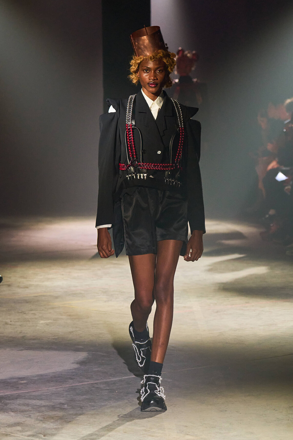 Model walks a fashion runway in a black tailored suit with red chains, metallic harness, and copper top hat.