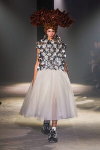 Model walks the runway in a bodice covered with oversized silver metallic flower appliqués and a white tulle midi skirt, paired with embellished boots.
