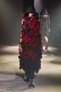 Runway model in a hooded black cloak covered with red and black flowers, paired with black pants and jeweled boots.