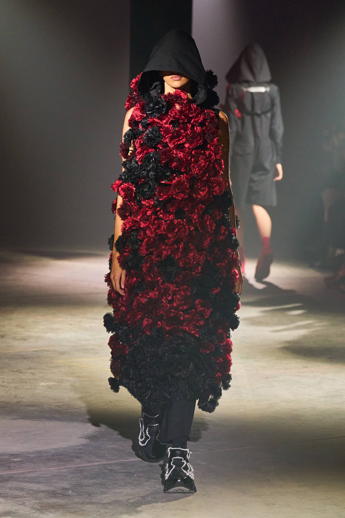 Runway model in a hooded black cloak covered with red and black flowers, paired with black pants and jeweled boots.
