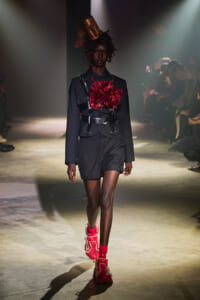 Model walks a runway in a black tailored suit with a large red satin bow and metallic harness, complemented by bright red sneakers under dramatic lighting.