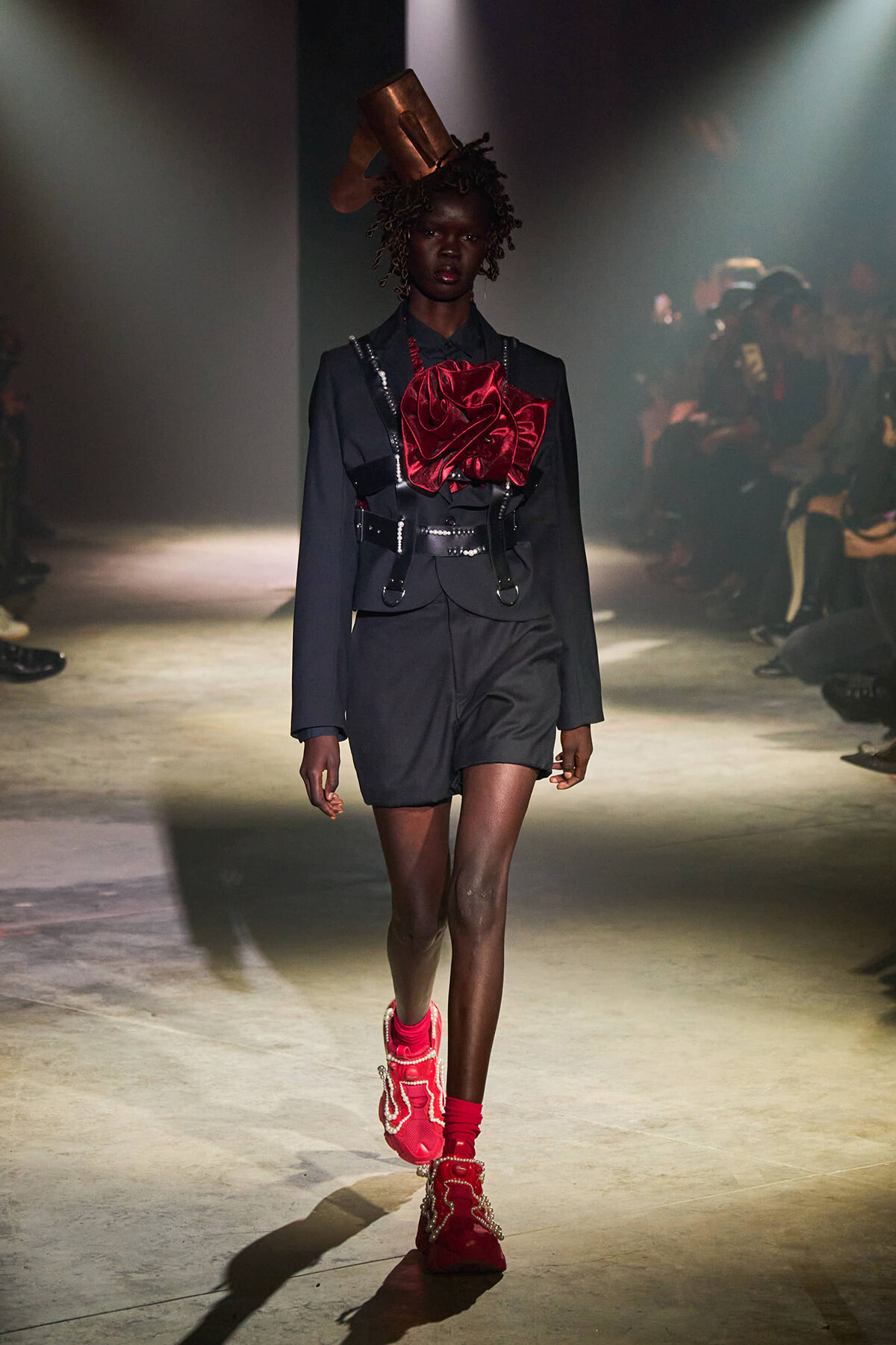 Model walks a runway in a black tailored suit with a large red satin bow and metallic harness, complemented by bright red sneakers under dramatic lighting.