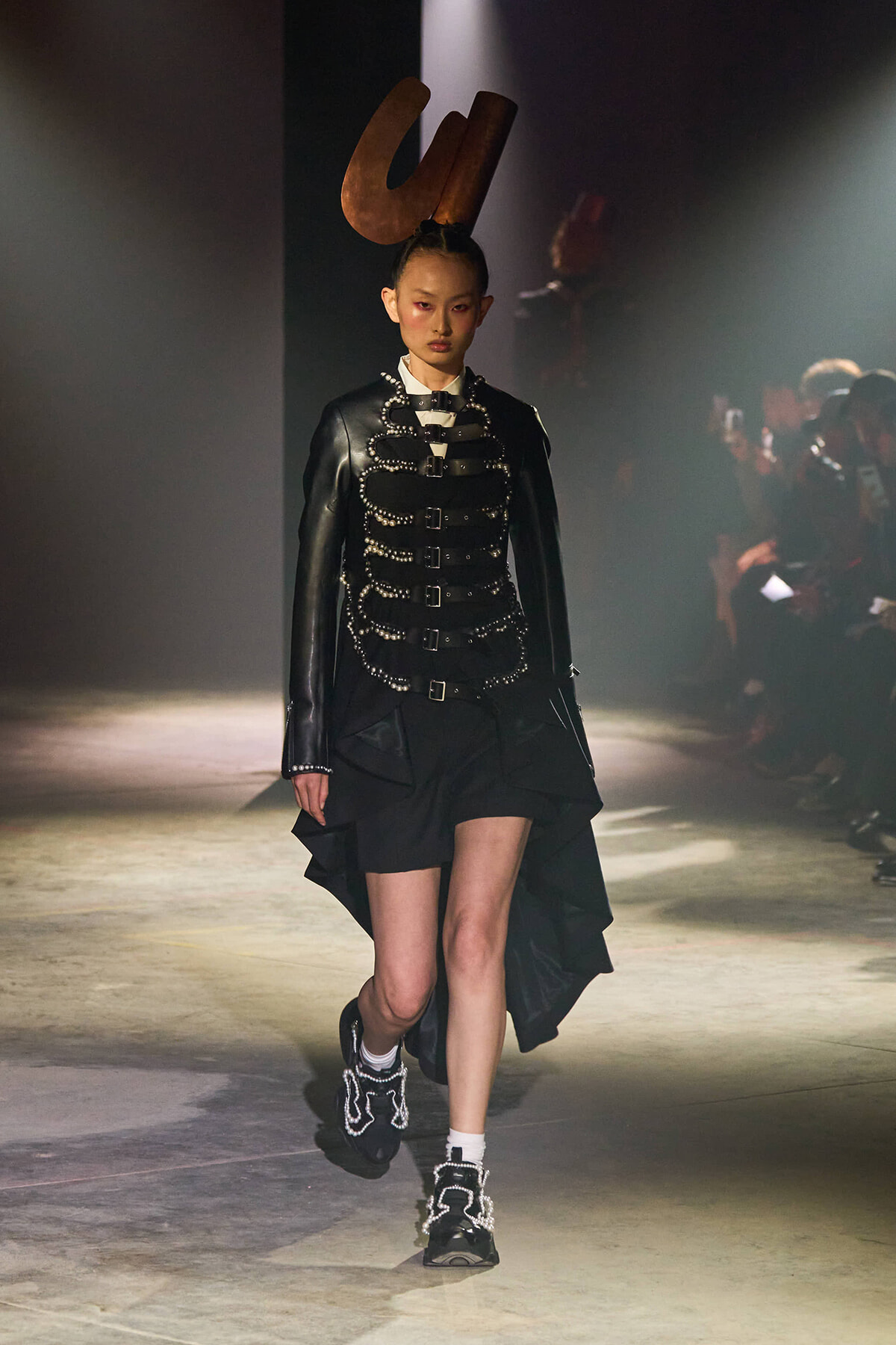 Runway model in a black leather jacket with buckled straps over a high-low skirt and a large brown U-shaped headpiece.