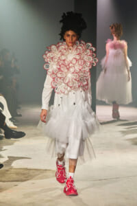 Model walking a runway in an avant-garde white ensemble with a giant pink-and-white floral collar and layered tulle skirt, pink sneakers visible in foreground.