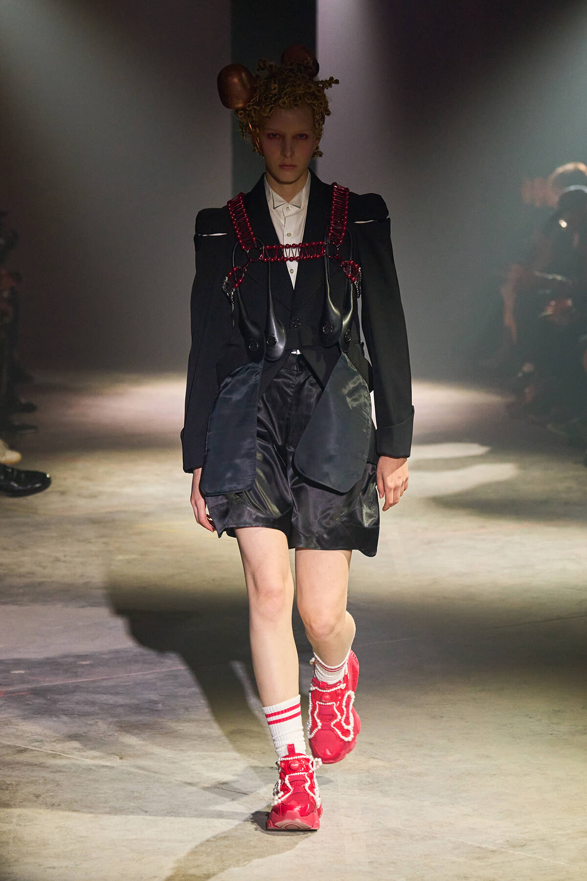 Model walks a fashion runway wearing a black blazer with a red harness and oversized pockets over satin shorts, paired with white socks and red sneakers.