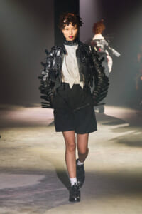 Model walks a runway in a futuristic black strappy armored jacket over a white shirt and black shorts, bold couture style.
