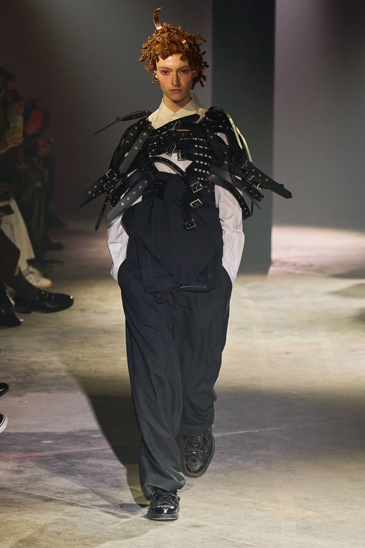 Model walks a runway wearing sculptural black strapped armor layered over a white shirt and dark jumpsuit, with red-toned hair.