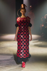 Model on a runway wearing a red circular-cutout lattice dress over black, with a copper sculptural hat and red sneakers under spotlight.